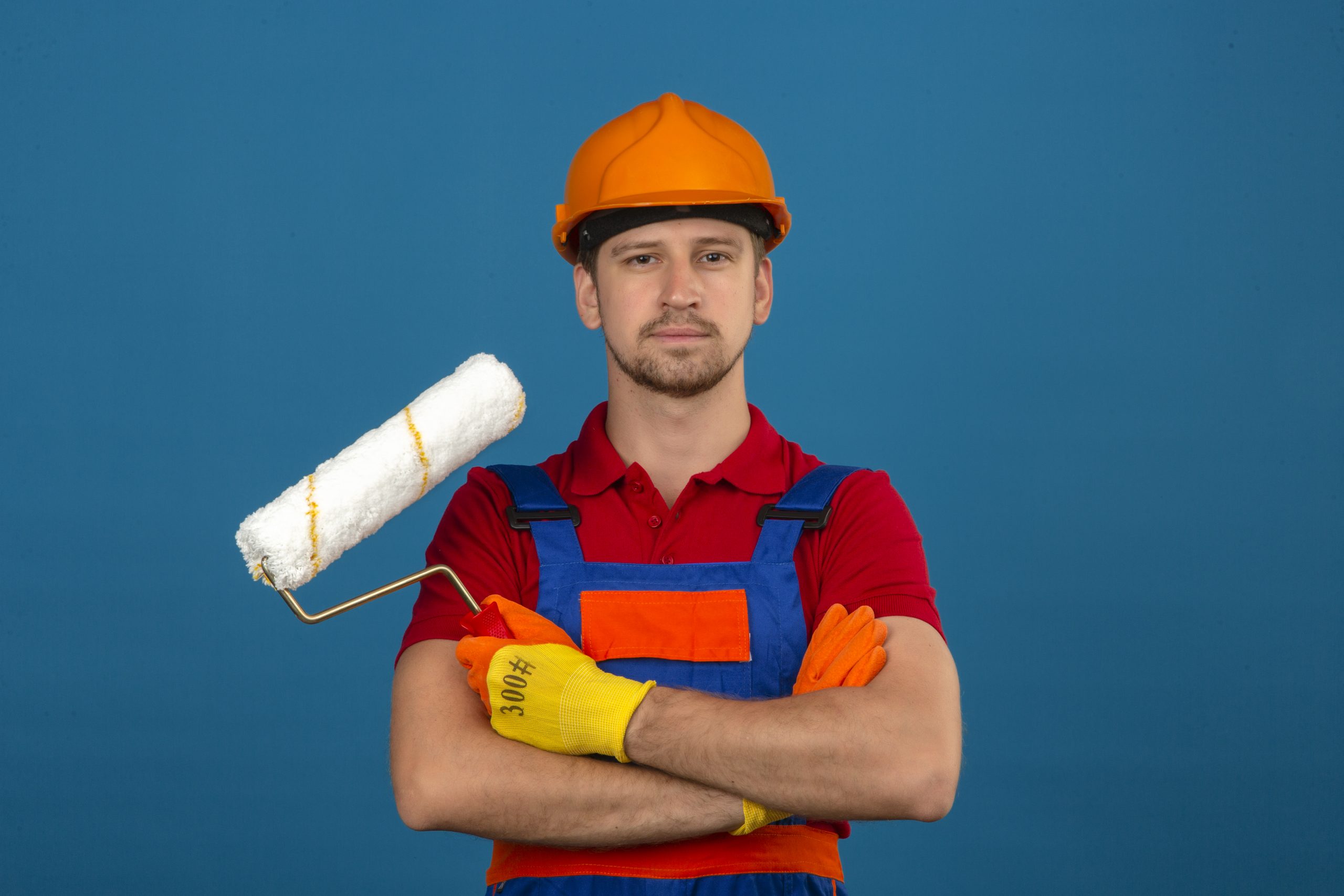 young builder man construction uniform safety helmet standing with crossed arms with paint roller looking confident isolated blue wall scaled-hayward-california